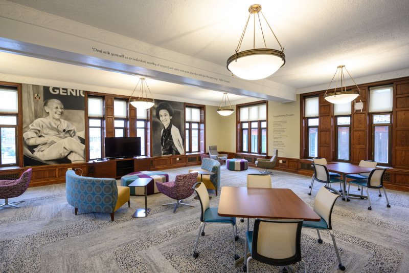 A brightly lit, modern lounge area with large windows and wood paneling. The room features comfortable, colorful seating with a mix of chairs, small tables, and ottomans arranged for casual meetings or relaxation.