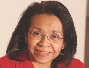 An image of Shirley Malcom smiling and wearing glasses in a red suit coat.