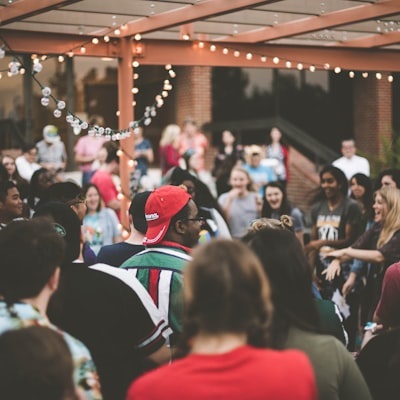 A lively outdoor gathering with a diverse group of people socializing under string lights. The event takes place in a courtyard or patio setting with a mix of casual and festive attire, creating a warm and celebratory atmosphere