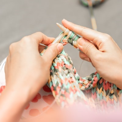 Close-up of hands knitting with thick, multicolored yarn made from recycled fabric, using wooden knitting needles. The background is blurred, and the person is wearing a light-colored outfit with red heart patterns, suggesting a focus on sustainable or zero-waste crafting.