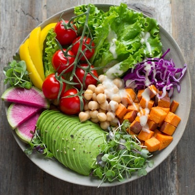 A colorful, plant-based salad bowl with fresh ingredients including sliced avocado, cherry tomatoes on the vine, chickpeas, roasted sweet potatoes, lettuce, shredded purple cabbage, yellow bell peppers, watermelon radish slices, and microgreens, drizzled with a light dressing. The bowl sits on a rustic wooden surface, emphasizing a fresh and healthy meal.