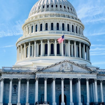 Photograph of the United States Capitol building with its iconic dome, columns, and an American flag flying in front. The clear blue sky serves as a backdrop, symbolizing governance, policy-making, and legislative discussions.