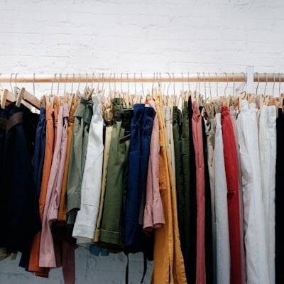 A clothing rack filled with a variety of colorful second-hand or sustainably sourced pants and trousers, neatly hung on wooden hangers against a white brick wall. The image suggests a focus on sustainable fashion, clothing swaps, or zero-waste shopping.