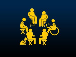 6 people sit in a circle on various chairs and assistive devices. 1 person has a service dog and another holds a laptop.