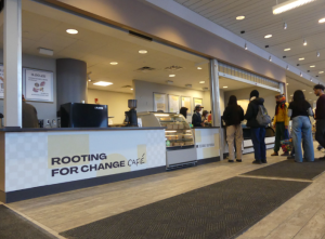 This image depicts a line of people waiting to order in front of a cafe counter. The counter reads "Rooting for Change Cafe."