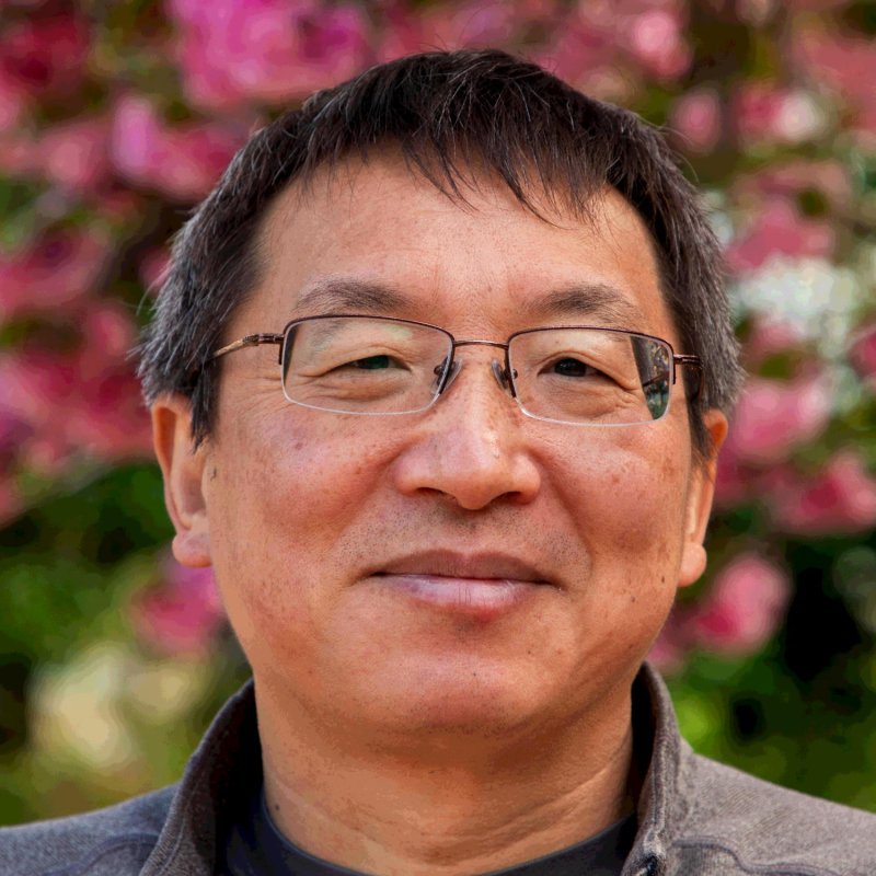 Headshot of Yong Cai. He has short dark hair, glasses, and is standing in front of pink florals.
