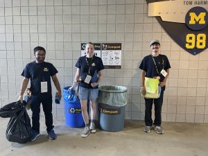 Three stadium volunteers stand in front of bin signage.