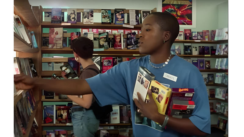 A person wearing a blue shirt and name tag browses a video rental store, holding several VHS tapes. Shelves filled with colorful movies surround them.