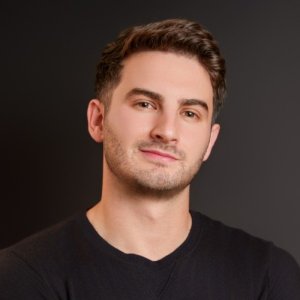 Brad Baum, a young man with short brown hair and light stubble, looks directly at the camera with a slight smile. He is wearing a black crewneck shirt against a dark, neutral studio background.