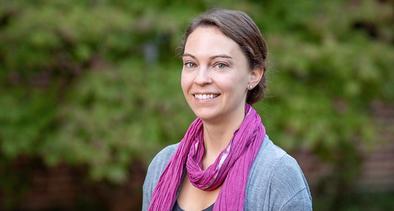 A person is standing outdoors in front of a background of green foliage. The person is wearing a grey cardigan over a dark shirt, accessorized with a bright pink scarf wrapped loosely around their neck. Their hair is pulled back, and they are wearing stud earrings. The setting appears to be calm and natural.