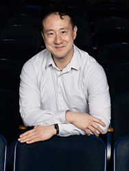 Edward Wang, in a white dress shirt, sitting in the audience area of a darkened theatre