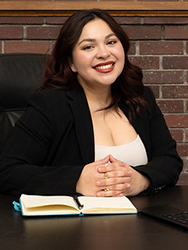 Isabella Valdez Martinez, dresses in a black jacket and a white top, sitting at a desk in front of a fireplace.