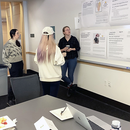 Three female students in the Impact Studio course looking at How-To poster on the wall