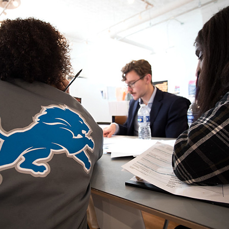 Black man in a Detroit Lions jacket with a pencil looks at young man looking at papers while a woman also looks on