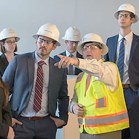 Construction engineer in hard hat points off camera as students in business students in hardhats and business suits look on