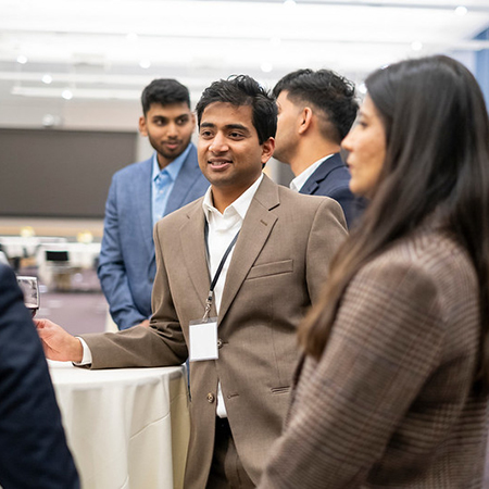 South Asian man smiles, speaking off camera, as diverse people stand in a group at a tall table