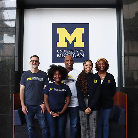 Diverse group of two men and three women standing in front of UM logo, wearing UM Detroit Center shirts of different colors and styles