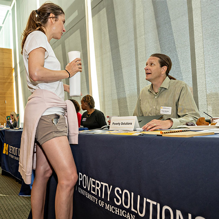 Young female student standing in front of exhibition table with older man, and the words Poverty Solutions on the table cloth