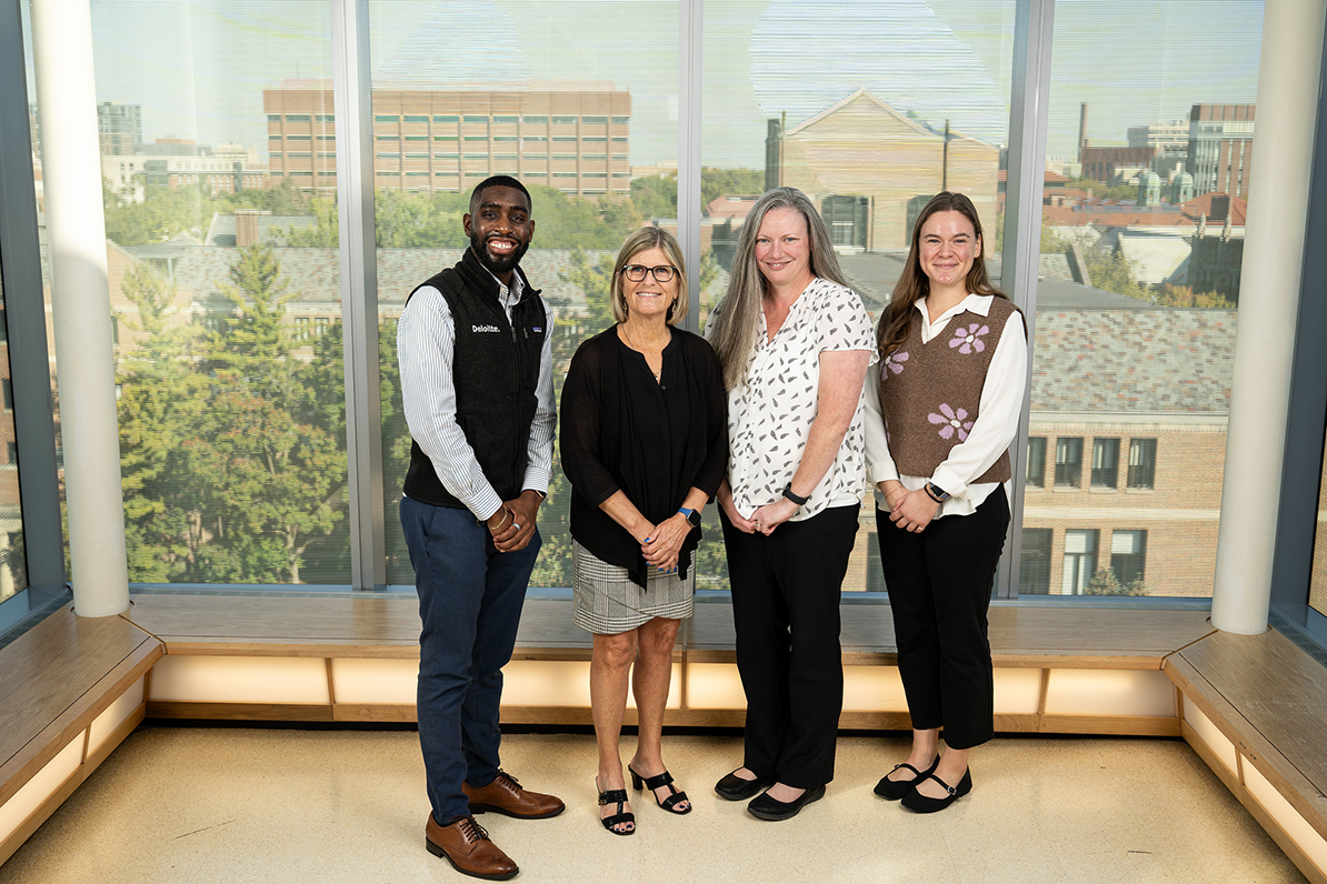 Black man, two white women and a white female student posted in front of a window with UM campus behind it