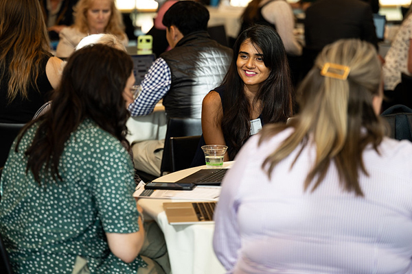Young woman of color smiling at another woman facing away, at a six-top table