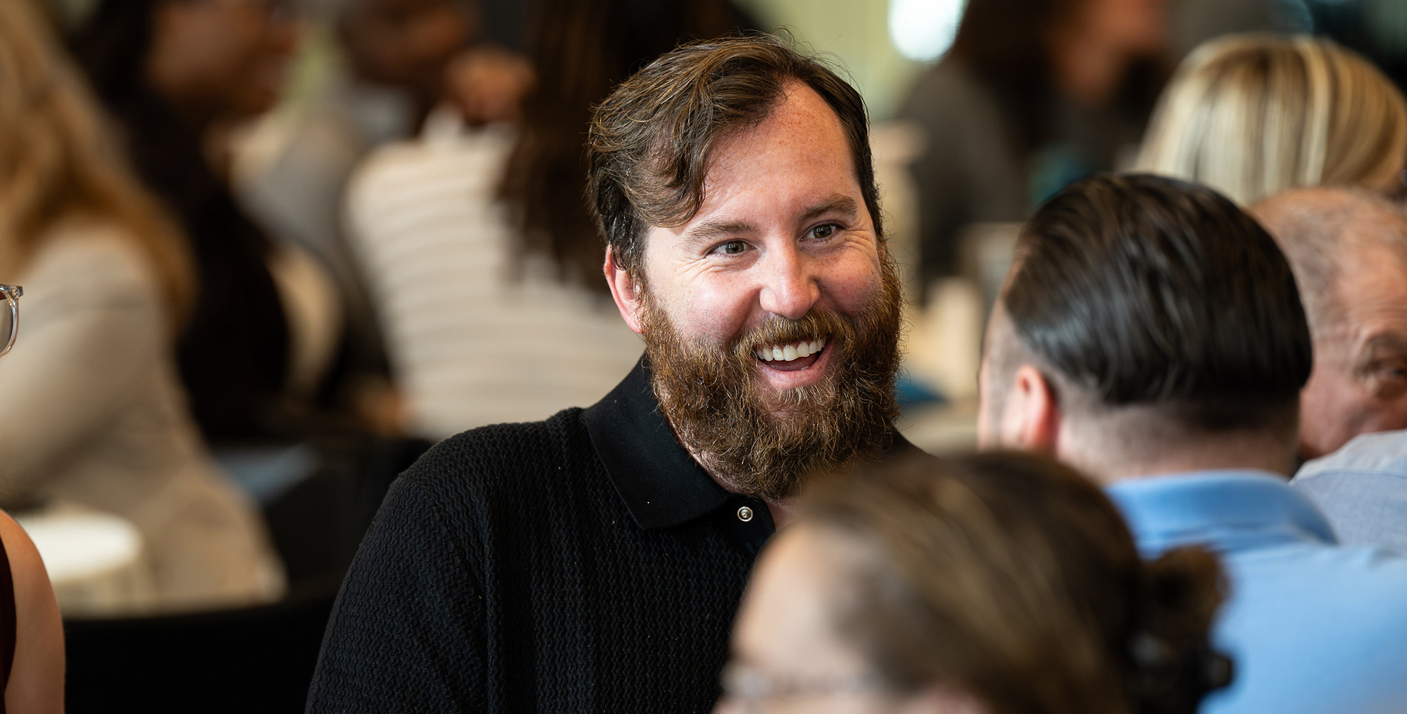 Bearded white man smilling, speaking to a tatooed white man, in a crowd of blurred out individuals