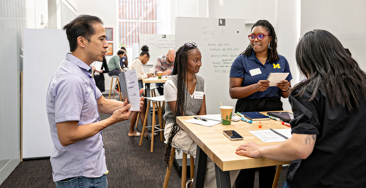 Moses Lee speaks to two black women in the Impact Studio as other diverse groups work in the background