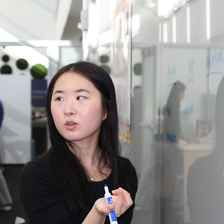 Asian woman holding marker in front of marker board, speaking to someone off screen
