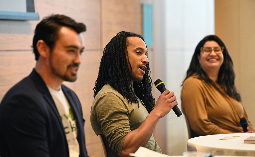 Black man with microphone speaking between a white man and latina sitting on a panel