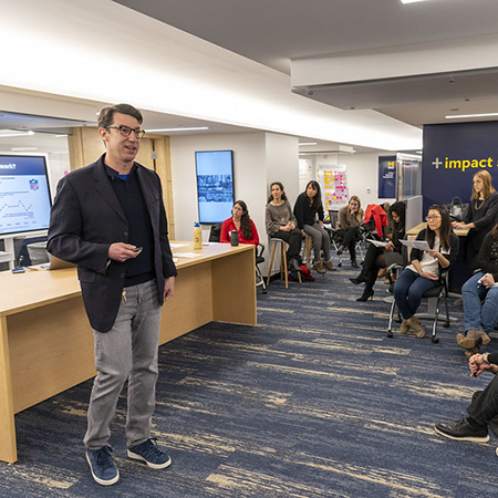 White man presenting ideas to a diverse crowd in the old Impact Studio