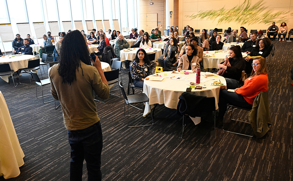 Adult with long hair speaking in front of a bunch of people at round tables.