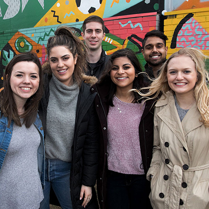 Diverse group of 6 students smiling at the camera in front of an outdoor mural