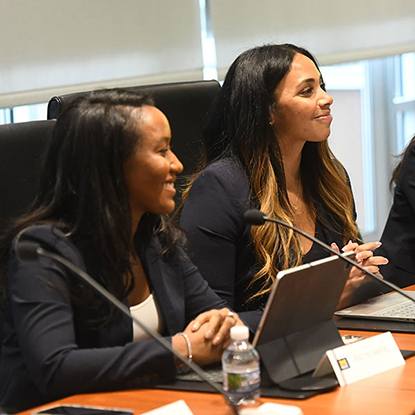 Black woman and Latina at a boardroom table, smiling toward the right