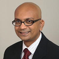 Shailesh Kumar in posed portrait, wearing a black suit with burgundy tie, and smiling