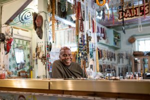 Artist Olayami Dabls behind a counter inside his bead museum filled with hanging beads, masks, and artwork.